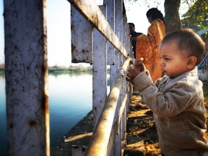 A young child with short hair wearing a sweater stands near a metal railing beside a calm body of water. The child is focused on something in their hands, possibly food. In the background, two adults, one draped in an orange shawl, are partially visible beside a tree under a clear sky.