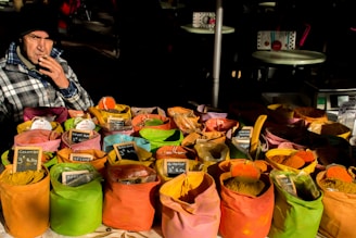 A man wearing a checkered jacket and hat is sitting behind a vibrant display of colorful spices at a market. Numerous cloth bags are filled with various spices, each labeled with small signs. The bags are arranged in neat rows, showcasing a variety of colors such as yellow, orange, pink, and green. The scene is set outdoors with a few tables in the background.