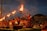 A group of men dressed in red traditional attire are performing a ritual with large lit oil lamps, creating a striking visual of flames against the evening sky. The ceremony is set in an outdoor space with an audience seated in the background, observing the ritual.