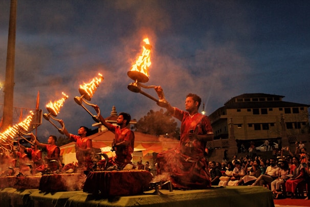 A group of men dressed in red traditional attire are performing a ritual with large lit oil lamps, creating a striking visual of flames against the evening sky. The ceremony is set in an outdoor space with an audience seated in the background, observing the ritual.