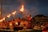A group of men dressed in red traditional attire are performing a ritual with large lit oil lamps, creating a striking visual of flames against the evening sky. The ceremony is set in an outdoor space with an audience seated in the background, observing the ritual.