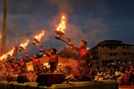 A group of men dressed in red traditional attire are performing a ritual with large lit oil lamps, creating a striking visual of flames against the evening sky. The ceremony is set in an outdoor space with an audience seated in the background, observing the ritual.