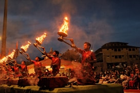A group of men dressed in red traditional attire are performing a ritual with large lit oil lamps, creating a striking visual of flames against the evening sky. The ceremony is set in an outdoor space with an audience seated in the background, observing the ritual.