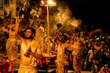 A group of people dressed in traditional attire are performing a religious ceremony involving the use of large brass lamps. Smoke is rising from the lamps, creating an atmospheric effect. The event is being watched by a large crowd, and some spectators are capturing the moment with their phones. The environment appears to be lively and spiritual, with a warm and inviting glow from the lamps.