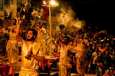 A group of people dressed in traditional attire are performing a religious ceremony involving the use of large brass lamps. Smoke is rising from the lamps, creating an atmospheric effect. The event is being watched by a large crowd, and some spectators are capturing the moment with their phones. The environment appears to be lively and spiritual, with a warm and inviting glow from the lamps.