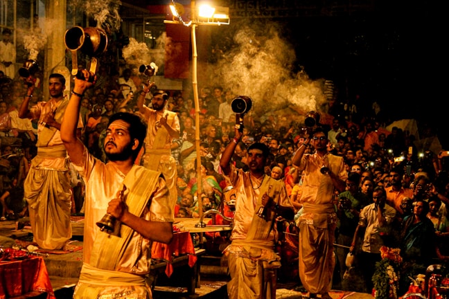 A group of people dressed in traditional attire are performing a religious ceremony involving the use of large brass lamps. Smoke is rising from the lamps, creating an atmospheric effect. The event is being watched by a large crowd, and some spectators are capturing the moment with their phones. The environment appears to be lively and spiritual, with a warm and inviting glow from the lamps.