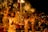A group of people dressed in traditional attire are performing a religious ceremony involving the use of large brass lamps. Smoke is rising from the lamps, creating an atmospheric effect. The event is being watched by a large crowd, and some spectators are capturing the moment with their phones. The environment appears to be lively and spiritual, with a warm and inviting glow from the lamps.