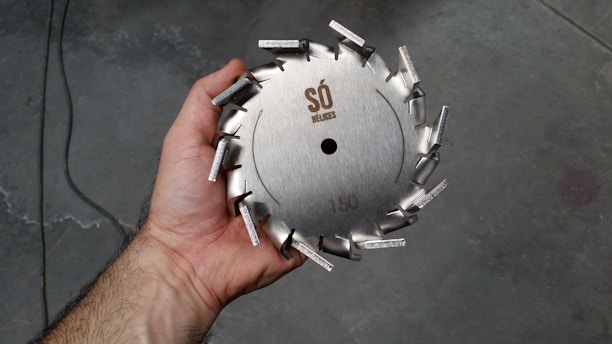 A hand holding a circular metal saw blade with protruding teeth arranged around the edge. The blade has engravings that include a logo and the number 150. The background appears to be a rough, industrial floor.