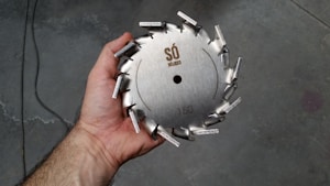 A hand holding a circular metal saw blade with protruding teeth arranged around the edge. The blade has engravings that include a logo and the number 150. The background appears to be a rough, industrial floor.