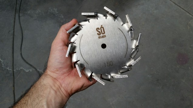 A hand holding a circular metal saw blade with protruding teeth arranged around the edge. The blade has engravings that include a logo and the number 150. The background appears to be a rough, industrial floor.