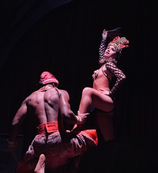 Two performers are on stage, with one standing confidently in an elaborate costume featuring a headdress and fishnet stockings, while the other is in a kneeling pose wearing a red outfit. The lighting is dim, creating a dramatic and theatrical atmosphere.