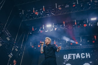 A performer passionately sings on stage, holding a microphone in front of a large LED screen with the word 'DETACH' visible. Stage lighting casts colorful beams, mostly red, with smoke effects enhancing the dramatic atmosphere. The stage setup includes numerous spotlights and rigging, with a crowd suggested at the bottom left corner.