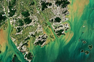 Aerial view of a coastal area with lush green vegetation, urban structures, and waterways interspersed. The region is bordered by a vibrant turquoise and brownish water body with small islands scattered around.