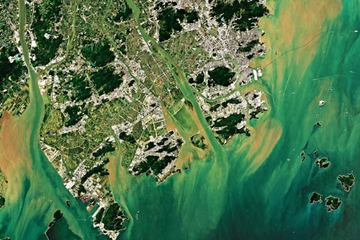 Aerial view of a coastal area with lush green vegetation, urban structures, and waterways interspersed. The region is bordered by a vibrant turquoise and brownish water body with small islands scattered around.