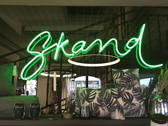 A vivid green neon sign spelling out the word 'Skand' is prominently displayed. In the foreground, decorative items including patterned pillows with leaf prints and two glass candle holders are arranged on a dark surface. The background features modern interior elements with metallic structures and lighting fixtures.