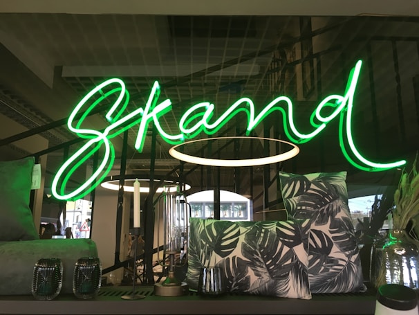 A vivid green neon sign spelling out the word 'Skand' is prominently displayed. In the foreground, decorative items including patterned pillows with leaf prints and two glass candle holders are arranged on a dark surface. The background features modern interior elements with metallic structures and lighting fixtures.