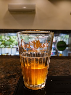 A clear glass with the brand 'Chai Kaale' written on it in orange, half-filled with an amber liquid. The glass is placed on a dark countertop with a blurred background showing a window through which trees and light are visible.