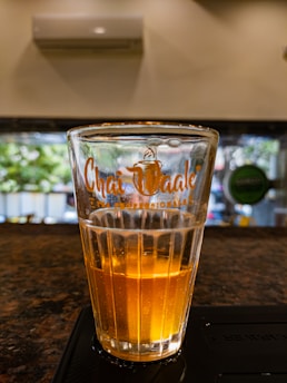 A clear glass with the brand 'Chai Kaale' written on it in orange, half-filled with an amber liquid. The glass is placed on a dark countertop with a blurred background showing a window through which trees and light are visible.