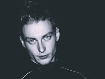 A monochrome portrait of a person with light skin, looking directly at the camera with a neutral expression. The background is dark, emphasizing the contrast with the light on the face. The person has high cheekbones, a furrowed brow, and hair pulled back.