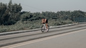 A cyclist is riding on an open road with greenery on the sides and a clear sky above. The cyclist is wearing a helmet and sportswear, appearing to be in a racing or training posture.