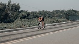 A cyclist is riding on an open road with greenery on the sides and a clear sky above. The cyclist is wearing a helmet and sportswear, appearing to be in a racing or training posture.