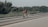 A cyclist is riding on an open road with greenery on the sides and a clear sky above. The cyclist is wearing a helmet and sportswear, appearing to be in a racing or training posture.