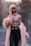 A person with long pink hair is wearing a stylish, sheer patterned top and a fluffy pink coat against a backdrop of softly blurred trees. The outfit is complemented by high-waisted dark pants with a black belt featuring a gold buckle.