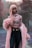 A person with long pink hair is wearing a stylish, sheer patterned top and a fluffy pink coat against a backdrop of softly blurred trees. The outfit is complemented by high-waisted dark pants with a black belt featuring a gold buckle.