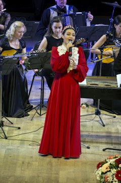 A female singer in a long red dress stands on a wooden stage, holding a microphone. She is surrounded by musicians playing instruments such as guitars and an accordion. The musicians are seated, and there are music stands in front of them with sheets of music. The stage is lit with bright lights, and there are flower arrangements on the floor.