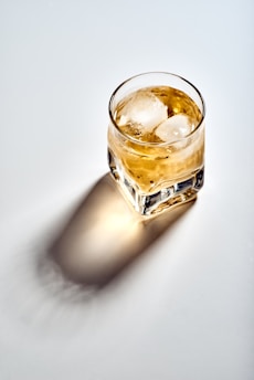 A glass filled with a caramel-colored beverage and ice cubes is positioned on a light surface, casting a soft shadow. The light source highlights the clarity of the glass and the texture of the ice.