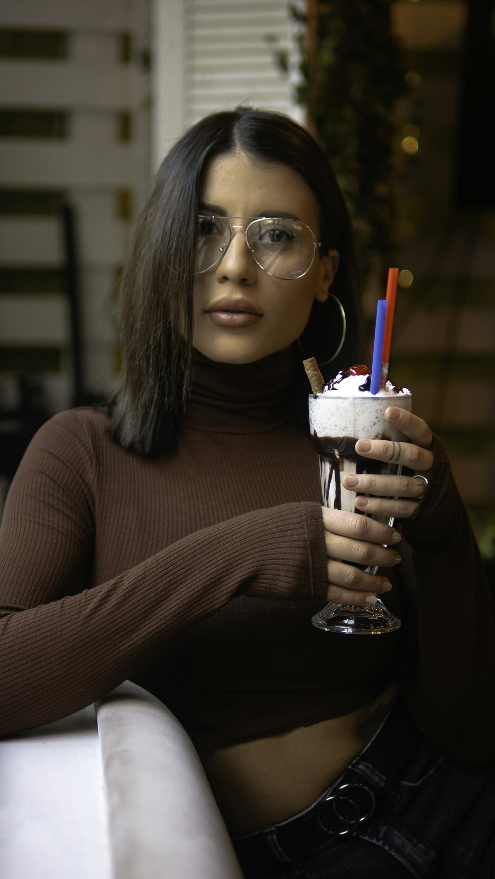 woman wearing brown turtleneck long-sleeved crop top holding cold drink while sitting on white chair