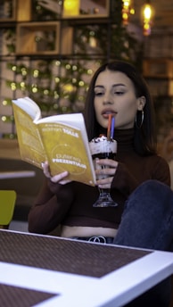 A person with long dark hair is sitting at a table, holding an open book with a yellow cover in one hand and a glass filled with a dessert topped with whipped cream in the other. The book's title is visible, and the person is sipping their drink through a straw. The background features shelves with plants and warm lighting.
