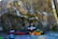 A person sits on a mat in front of a large, textured rock face, suggesting an outdoor climbing setting. The individual appears relaxed, surrounded by various climbing gear including pads and a backpack. Sunlight filters through the trees, creating spots of light on the rock surface.