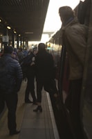 Commuters congregate on a dimly lit train platform, engaging in conversations and preparing to board or disembark from a train. The platform is crowded with people, and there is a sense of movement as one person is stepping onto the train. The setting sun casts a warm glow over the scene.