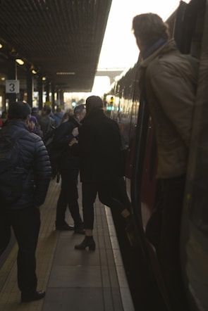Commuters congregate on a dimly lit train platform, engaging in conversations and preparing to board or disembark from a train. The platform is crowded with people, and there is a sense of movement as one person is stepping onto the train. The setting sun casts a warm glow over the scene.