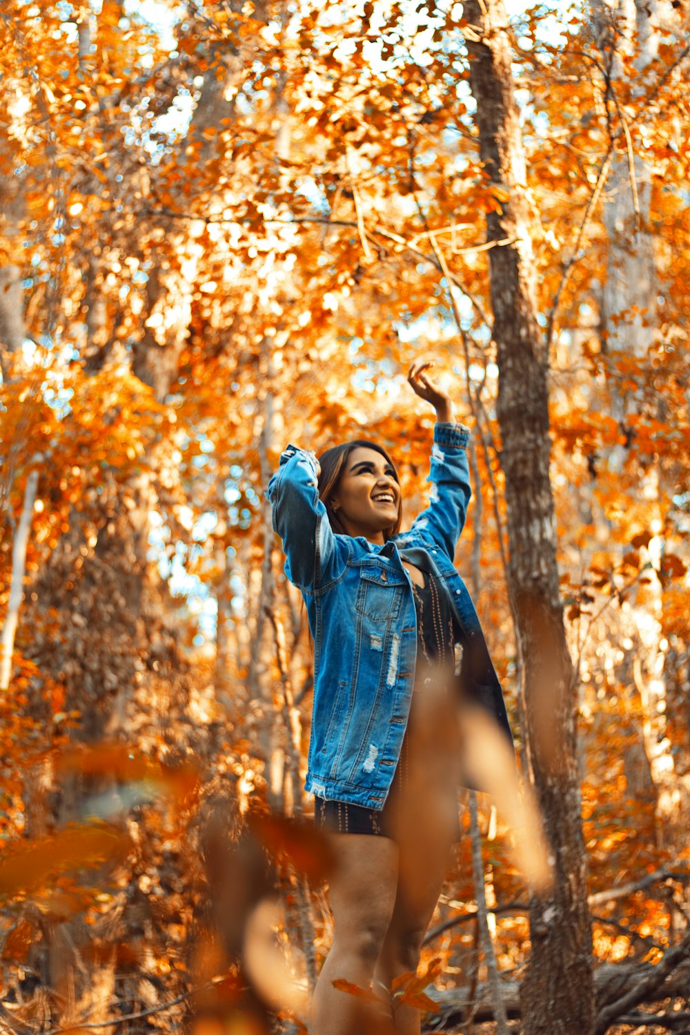 smiling woman wearing blue denim jacket standing while raising both hands surrounded with orange trees during daytime