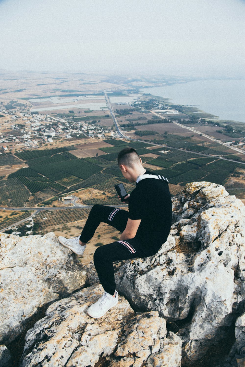 man wearing black t-shirt using phone while sitting on rocky hill viewing houses on green field during daytime