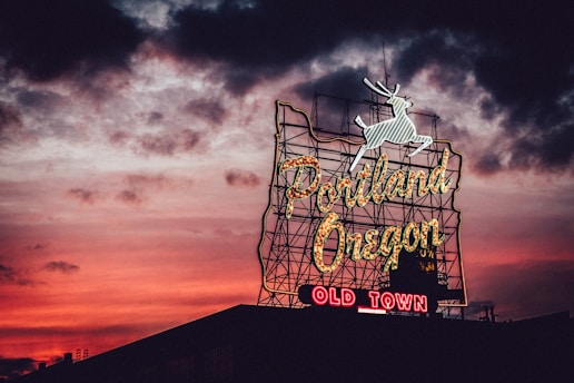 A neon sign featuring the words 'Portland Oregon' with 'Old Town' below, illuminated against a dramatic sunset sky. The sign is topped by a white stag silhouette, and the sky is a vibrant blend of red, orange, and dark clouds.