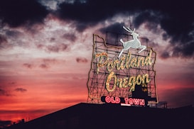 A neon sign featuring the words 'Portland Oregon' with 'Old Town' below, illuminated against a dramatic sunset sky. The sign is topped by a white stag silhouette, and the sky is a vibrant blend of red, orange, and dark clouds.