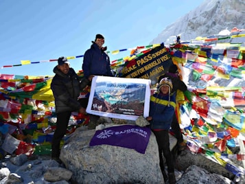 Three individuals stand beside a cairn at a high-altitude pass, surrounded by colorful prayer flags. They hold a banner with text about a 14-day journey and travel company information. The backdrop features snow-covered mountains under a clear blue sky.