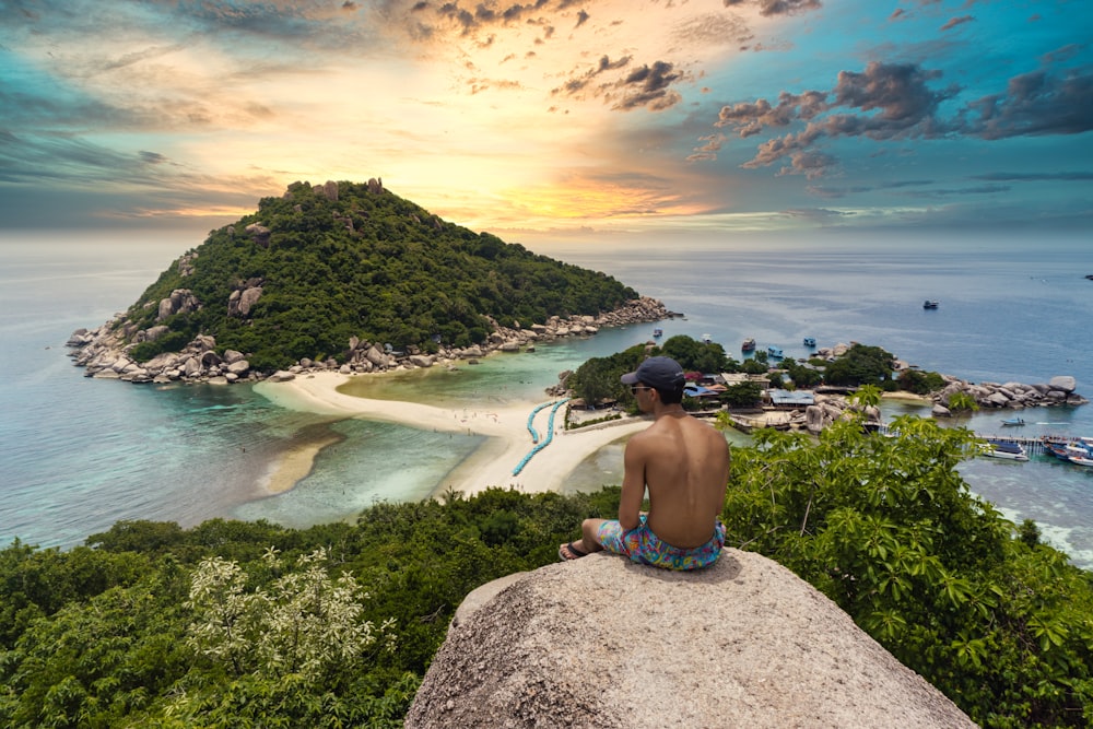 topless man sitting on cliff rock viewing Ko Nang Yuan isalnd, houses, and buildings under white and yellow sky