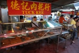 A bustling market scene featuring a meat vendor with a red and yellow sign marked 'Tuck Kee'. The vendor is behind a glass display case filled with various types of meat products, including square slices and sausages. Several people are seen shopping and engaging in transactions, with goods visibly stacked in the background.