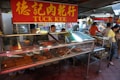 A bustling market scene featuring a meat vendor with a red and yellow sign marked 'Tuck Kee'. The vendor is behind a glass display case filled with various types of meat products, including square slices and sausages. Several people are seen shopping and engaging in transactions, with goods visibly stacked in the background.