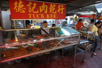 A bustling market scene featuring a meat vendor with a red and yellow sign marked 'Tuck Kee'. The vendor is behind a glass display case filled with various types of meat products, including square slices and sausages. Several people are seen shopping and engaging in transactions, with goods visibly stacked in the background.