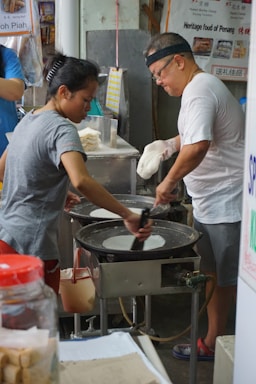 Two individuals are engaged in preparing food at a street market stall. One person is spreading batter on a hot griddle while the other is holding a tool to assist in the cooking process. There are various kitchen utensils and containers around them, and signage in the background indicates a focus on traditional food.