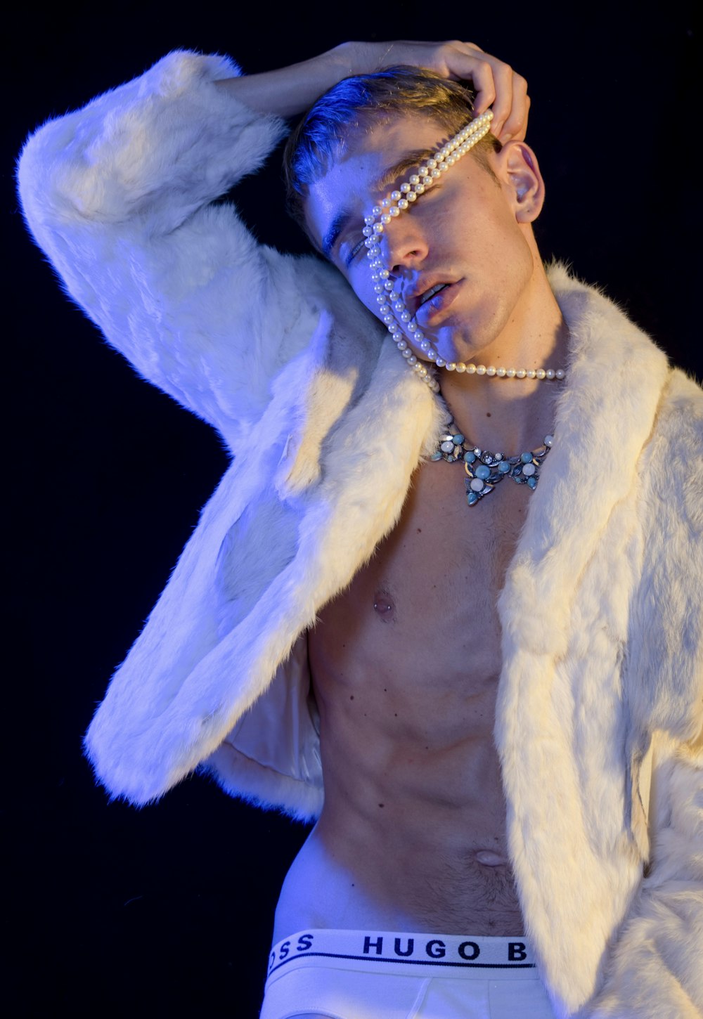 man wearing white fur jacket and white Hugo Boss brief standing while putting white pearl necklace on his head