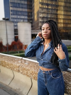 A person with long braided hair is dressed in a denim jacket and jeans, standing outdoors in an urban setting. Behind them is a background of modern glass buildings and concrete structures, with some foliage visible.