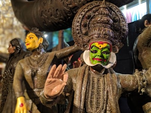 Intricately carved wooden sculptures depicting traditional theater characters with vibrant facial painting. The central figure has a green face with a dramatic headdress, wearing ornamental attire. Surrounding figures exhibit similar craftsmanship, highlighting cultural artistry.