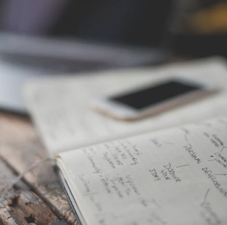 An open notebook with handwritten notes is placed on a wooden table next to a smartphone. The notes appear to focus on personal development and creative values. A partially visible laptop is in the background, creating a workspace setting.