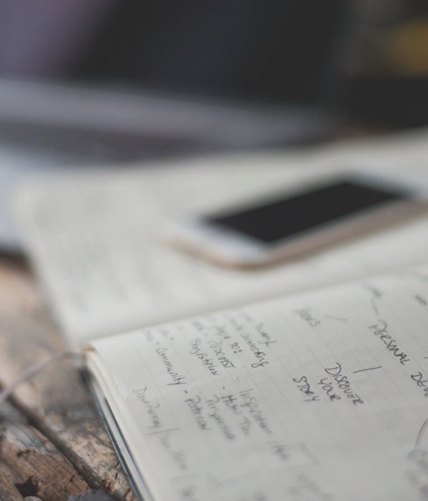 An open notebook with handwritten notes is placed on a wooden table next to a smartphone. The notes appear to focus on personal development and creative values. A partially visible laptop is in the background, creating a workspace setting.